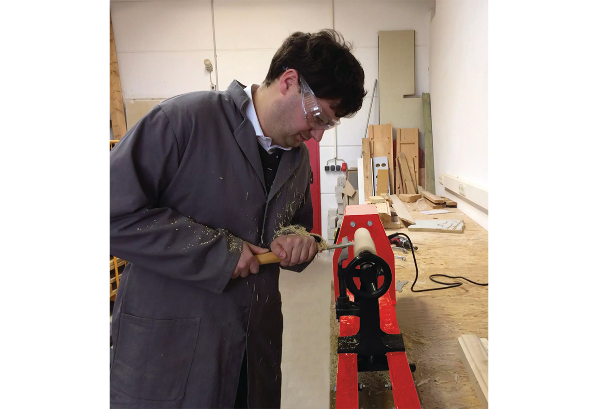 Απεικονίζεται άνδρας με προστατευτικά γυαλιά να σμιλεύει, χρησιμοποιόντας τον ξυλότορνο.